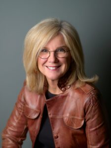 A woman with shoulder-length blonde hair and glasses is smiling. She is wearing a brown leather jacket and a black top, set against a plain grey background.