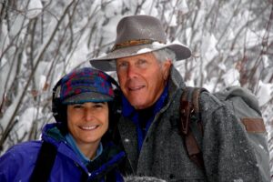 Two people in winter clothing smile at the camera, standing outdoors with snowy trees in the background.