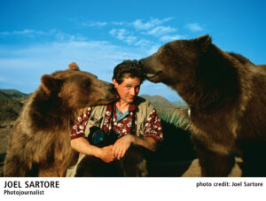 A person sits between two brown bears in an outdoor setting, one bear nuzzling his ear and the other bear licking his head. The person holds a camera.