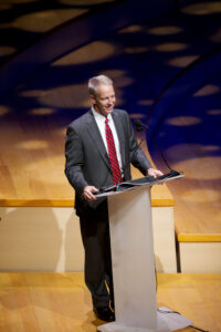 A man wearing a gray suit and red striped tie speaks at a podium on a wooden stage.