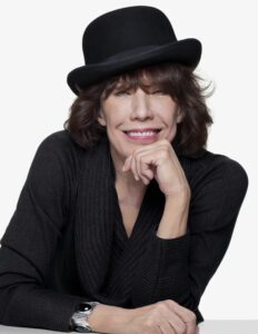 A person with medium-length brown hair smiles, wearing a black hat and a dark sweater, while resting their chin on their hand.