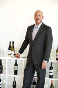 A bald man in a pinstripe suit stands by a shelf displaying assorted wine bottles and wine glasses.