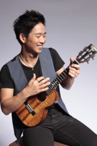 A person with short black hair and a black shirt under a gray vest is smiling and playing a ukulele against a plain background.