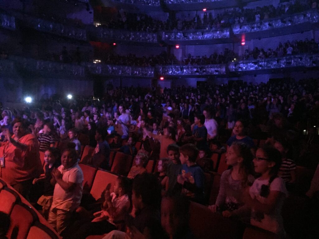 A dimly lit theater filled with a large audience, including many children, some standing and others seated, with a focus on the lower level. The upper balconies are also occupied.