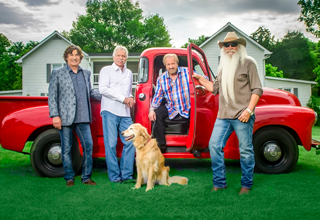 Four men and a dog pose beside a red vintage pickup truck in a grassy yard with houses and trees in the background. One man stands with the driver's door open.