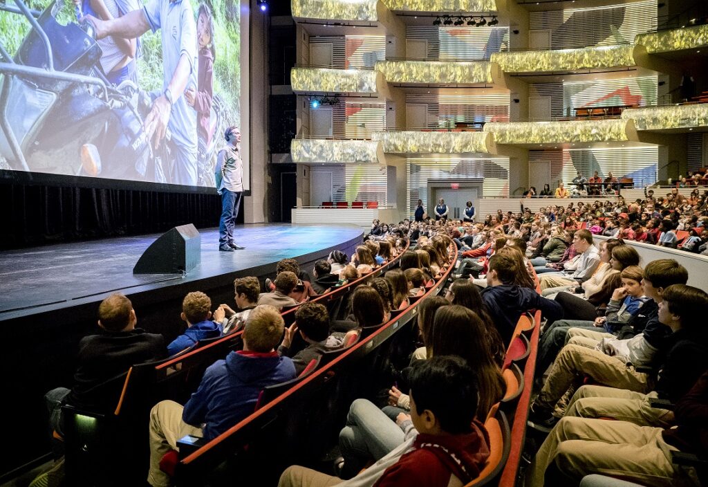 A speaker stands on a stage addressing a large audience seated in theater-style seating. A large screen displays an image behind the speaker.
