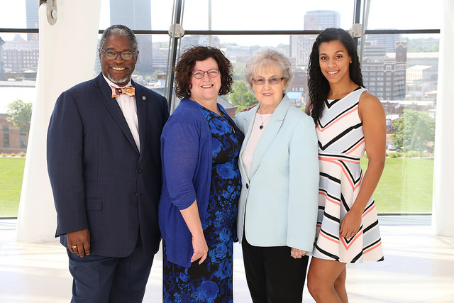 Four people stand side by side indoors with a cityscape visible through large windows in the background. They are dressed in formal and semi-formal attire, smiling at the camera.