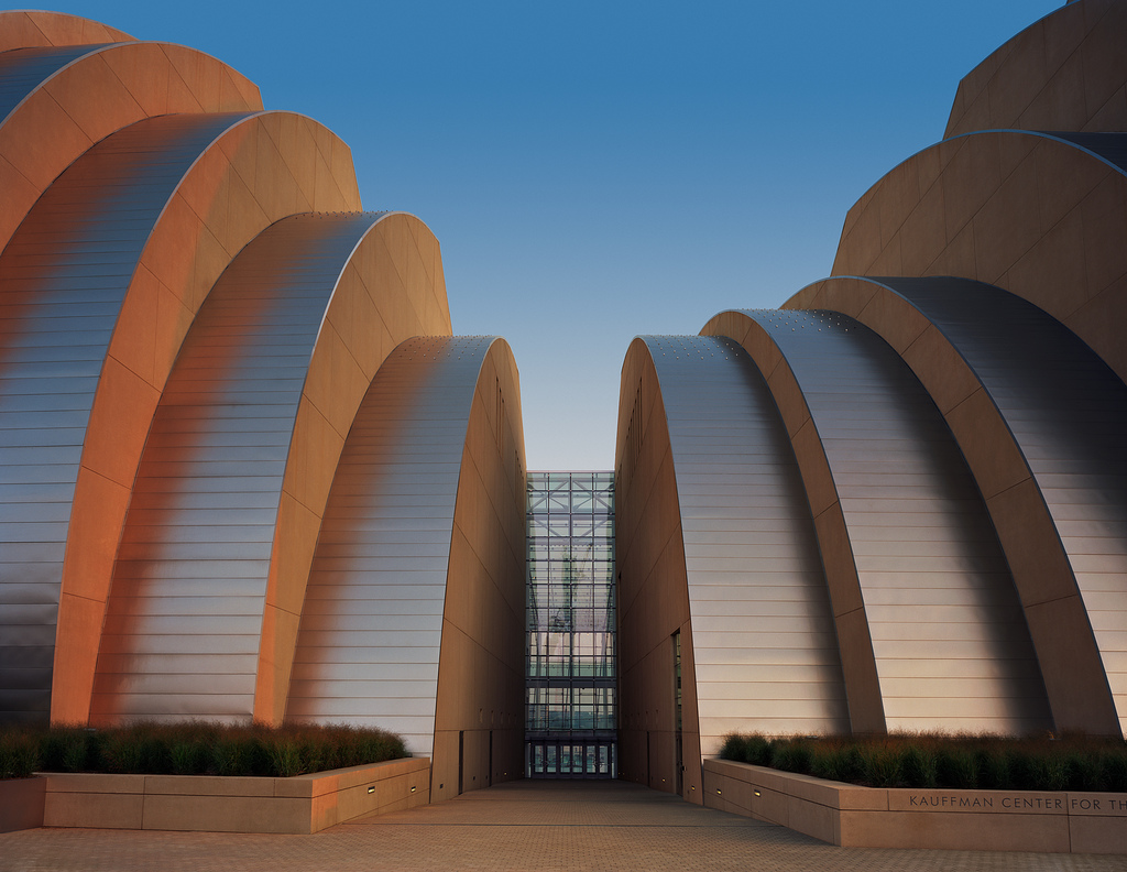 The image shows the Kauffman Center for the Performing Arts, a modern architectural building with curved, metallic surfaces and large glass windows, lit by the setting sun.