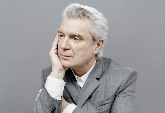 A person with gray hair wearing a gray suit rests their chin on their hand against a plain background.