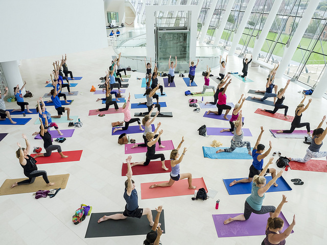 A large group of people practice yoga indoors, performing warrior poses on mats in a bright, spacious room with large, curved windows.