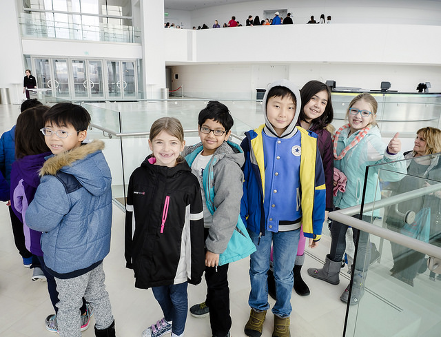 A group of children wearing winter clothes stand in line at an indoor location with glass railings and white walls. Some children are smiling at the camera while others look ahead.