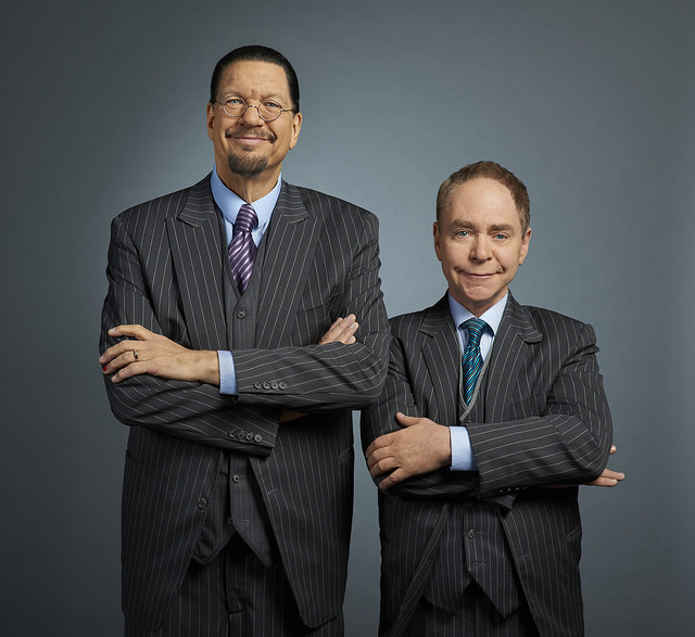 Two men wearing striped suits stand side by side against a gray background, both with arms crossed and smiling.