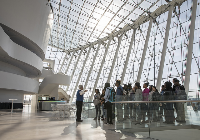 A group of people stands near a guide inside a modern building with large, curved architectural features and floor-to-ceiling windows.