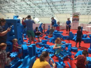 Children and adults are engaged in play with large blue foam building blocks on a red floor in a glass-enclosed space.