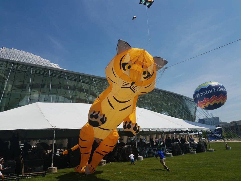 A large tiger-shaped kite is flying in front of a glass building with a white tent and crowd beneath it. A smaller kite and balloon with "Saint Luke's" written on it are also visible in the sky.