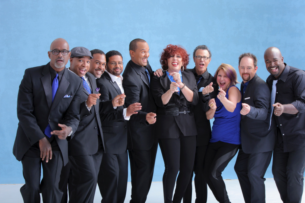 A group of ten people, dressed in formal and casual attire, stand closely together against a blue backdrop, smiling and making cheerful gestures.