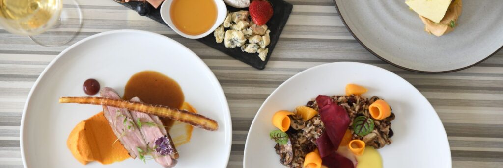 A table with three elegantly plated dishes featuring meat, vegetables, sauce, and garnishes, plus a small slate with nuts and a bowl of sauce. A glass of white wine is visible in the corner.
