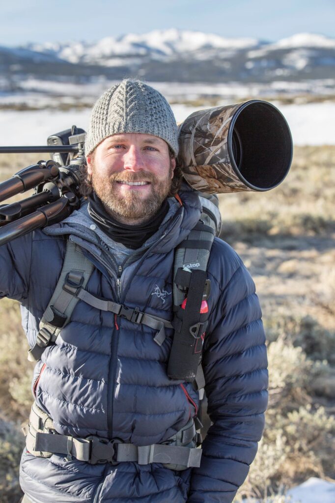 A person wearing a gray beanie and blue jacket smiles at the camera while carrying a large, camouflaged lens on their shoulder against a snowy mountain backdrop.