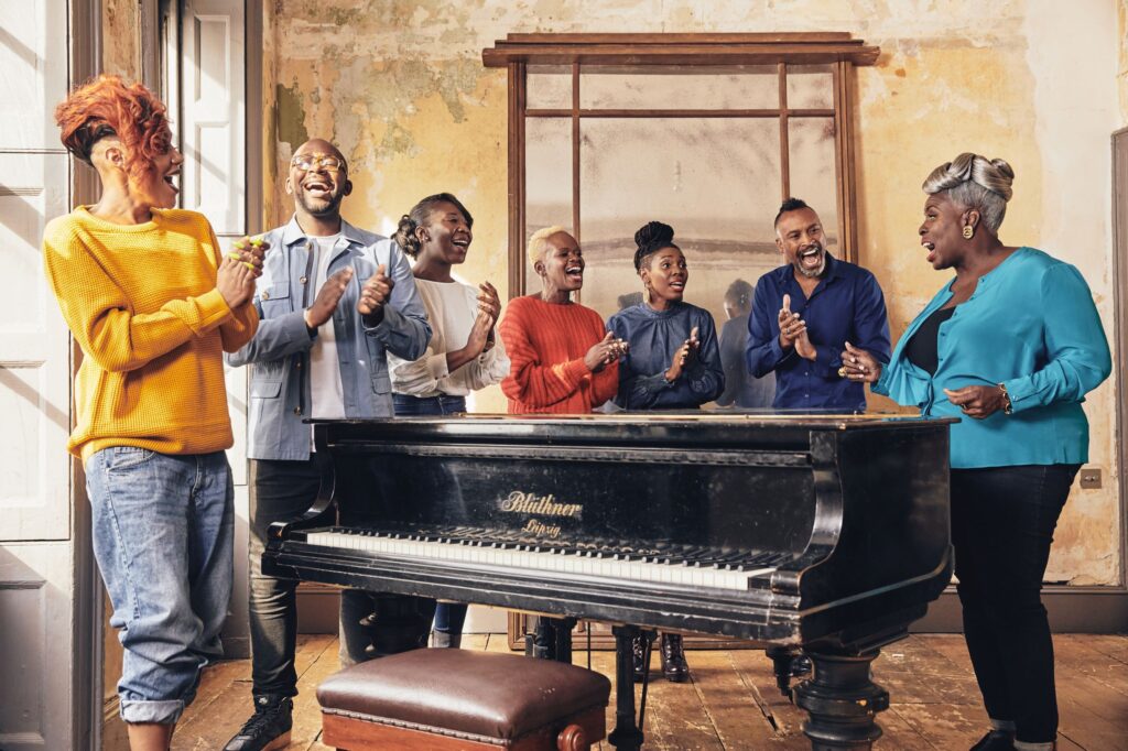A group of people standing around a black piano, singing and clapping in a warmly lit room with weathered walls and a wooden floor.
