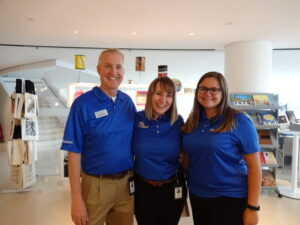 Three people wearing blue shirts and name tags stand together and smile in a well-lit room with shelves of books and merchandise in the background.