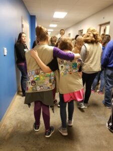 Two girls in uniform vests adorned with patches walk down a hallway with their arms around each other, surrounded by a group of people.