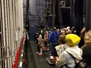 A group of people is touring a backstage area, observing the rigging system and stage equipment. One person is explaining.