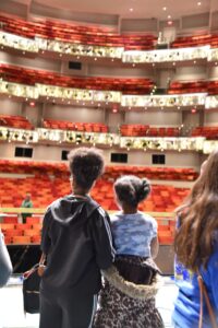 Two individuals with their backs to the camera facing an empty, multi-tiered theater with red and orange seats.