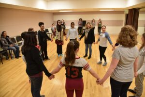 A group of people stand in a circle holding hands in a room. Some are standing, while others are seated along the walls. They appear to be participating in a group activity.