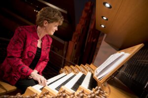 A person in a red jacket plays a large pipe organ, reading sheet music under overhead lights.