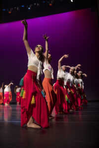 Dancers in vibrant costumes perform a choreographed routine on a dimly lit stage with a purple backdrop.