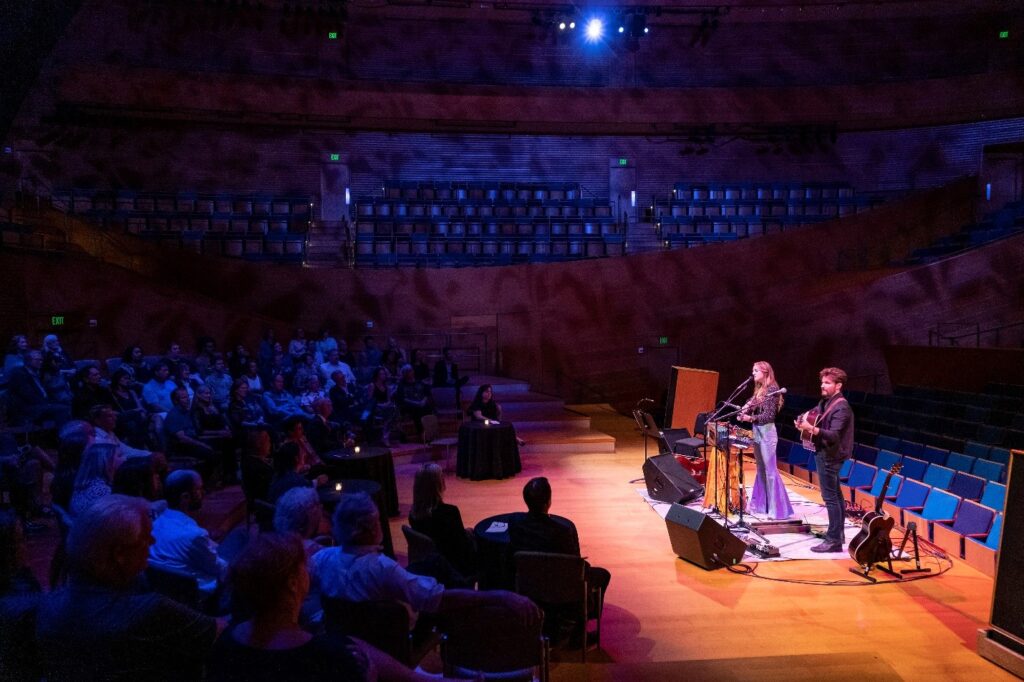 A duet performs on stage in a dimly lit concert hall with audience members seated in front of them. The background consists of empty seats extending to the upper levels.