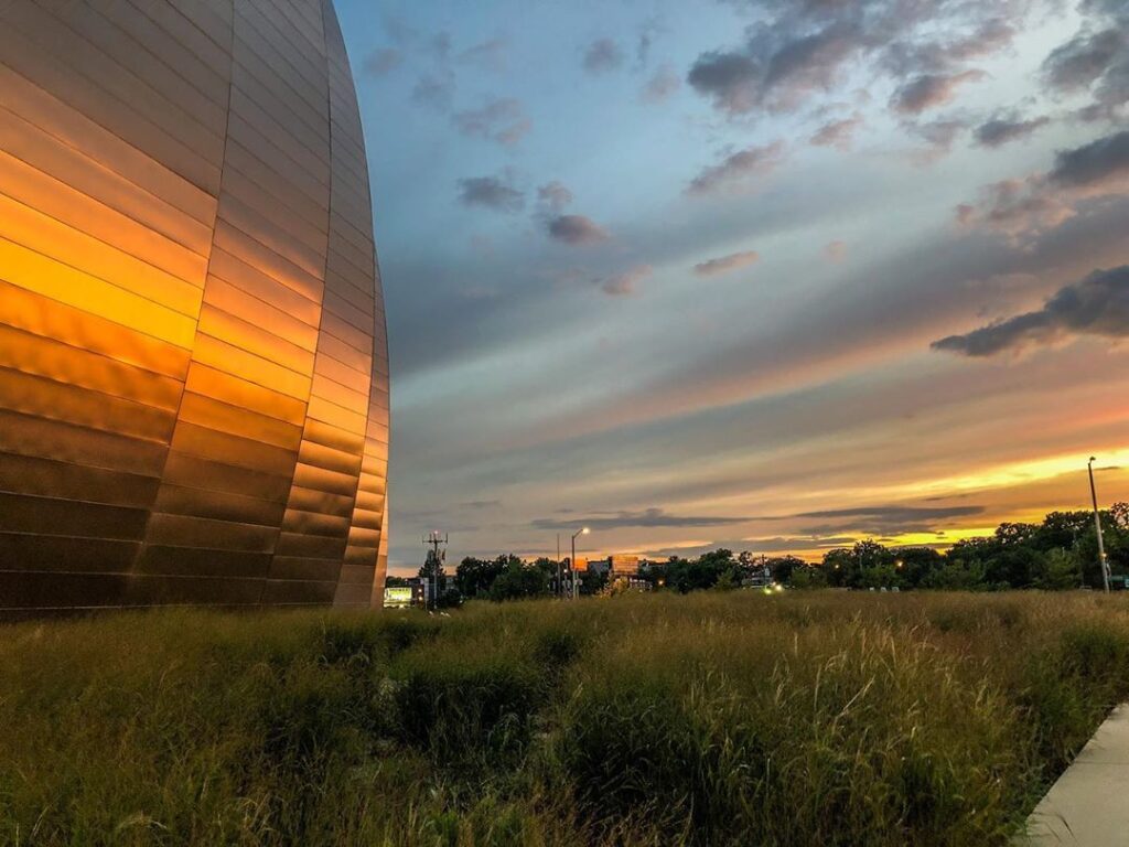 A curved metal building reflects the sunset, with grass and trees in the foreground and a colorful sky with clouds in the background.