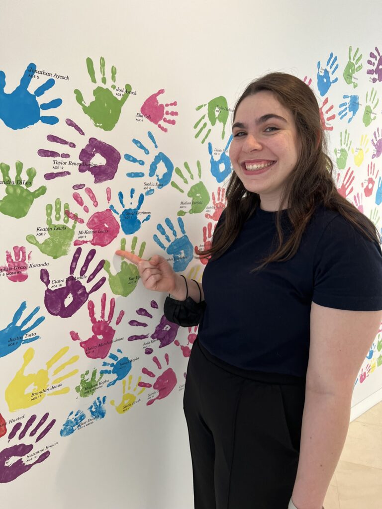 A woman in a black shirt stands in front of a wall covered with colorful handprints, smiling and pointing at a green handprint.