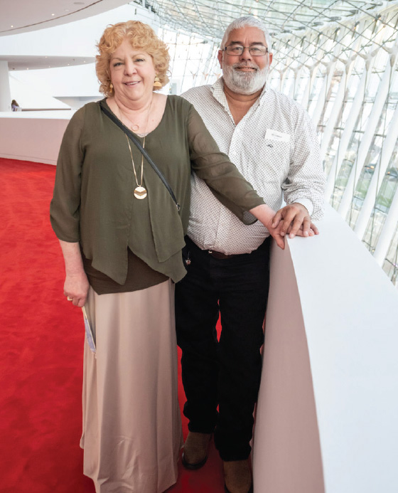 A woman and a man standing together in a modern building with a glass roof, leaning on a white railing with a red carpeted floor.