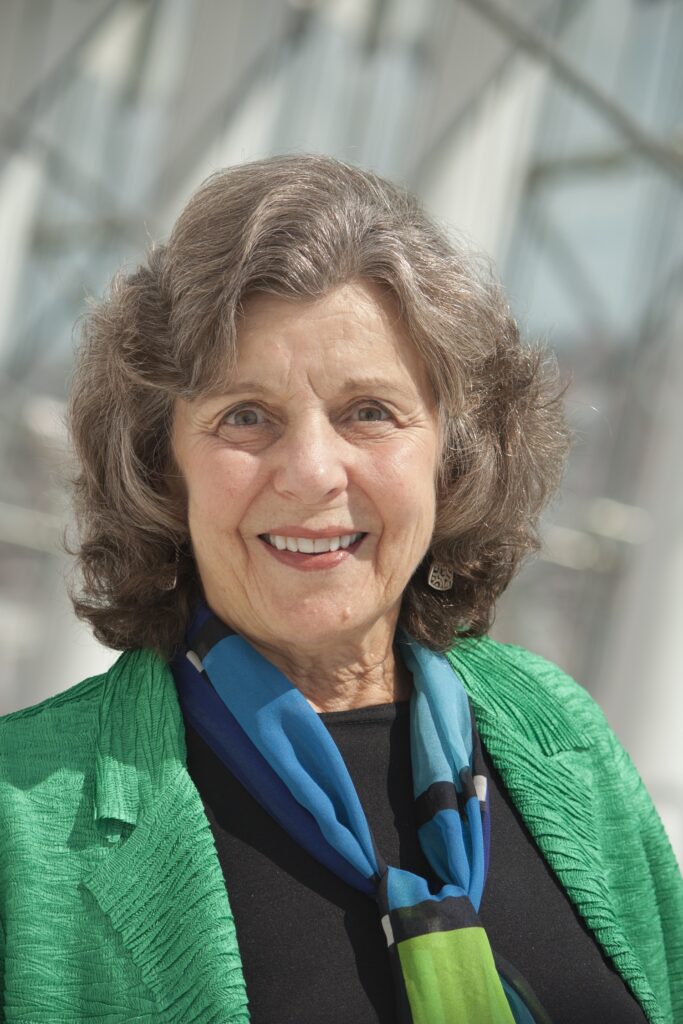 An older woman with wavy gray hair, wearing a green jacket and a blue scarf, smiles at the camera.