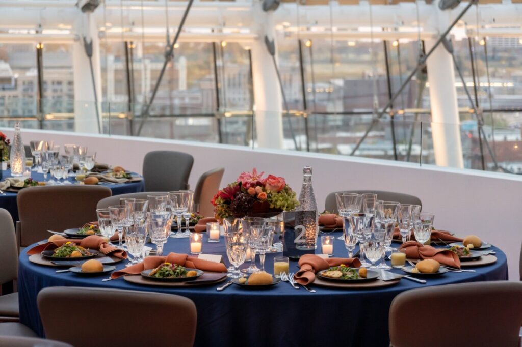 Elegant dining tables set for an event with blue tablecloths, folded napkins, glasses, cutlery, floral centerpieces, and candles. The background shows a cityscape through large windows.