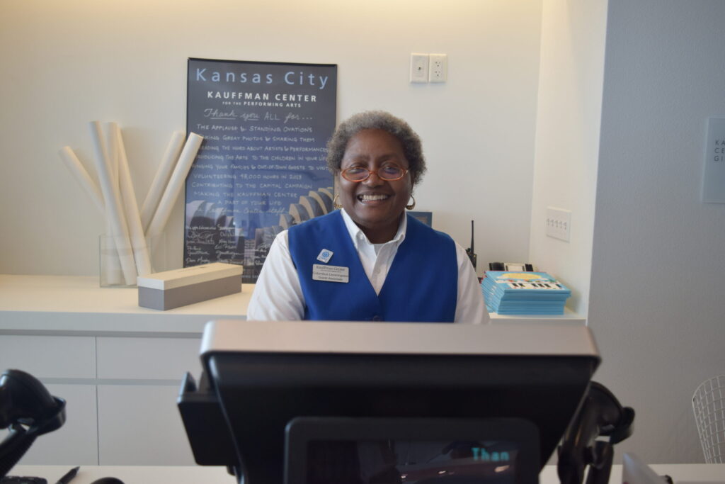 A person wearing a blue vest and name badge is standing behind a reception desk at the Kauffman Center in Kansas City, smiling at the camera.