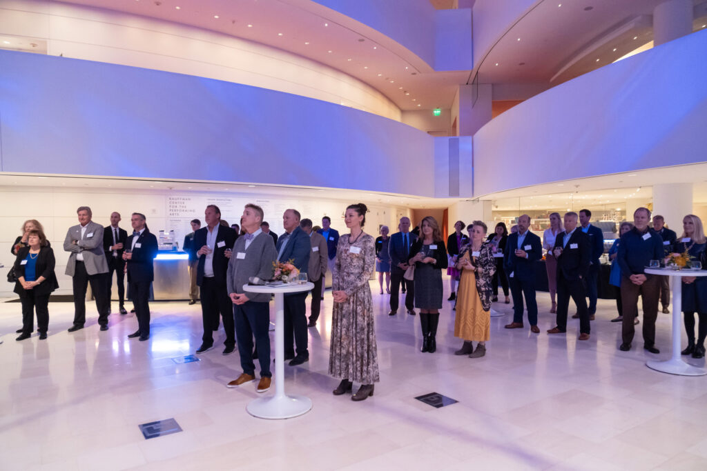 A group of individuals stands in a well-lit, modern hall with curved architectural features, gathered around tall tables. The setting appears to be a formal or corporate event.