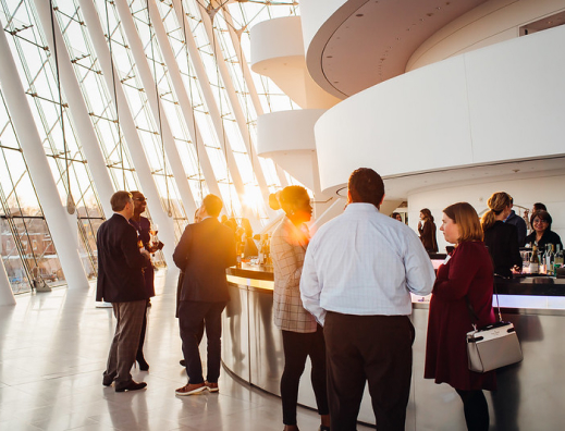People socializing in a modern atrium with floor-to-ceiling windows and curved architectural features as the sun sets.