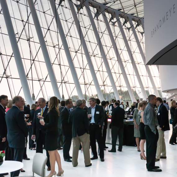 A large group of people in business attire socialize at an indoor event with tall windows and a modern design.