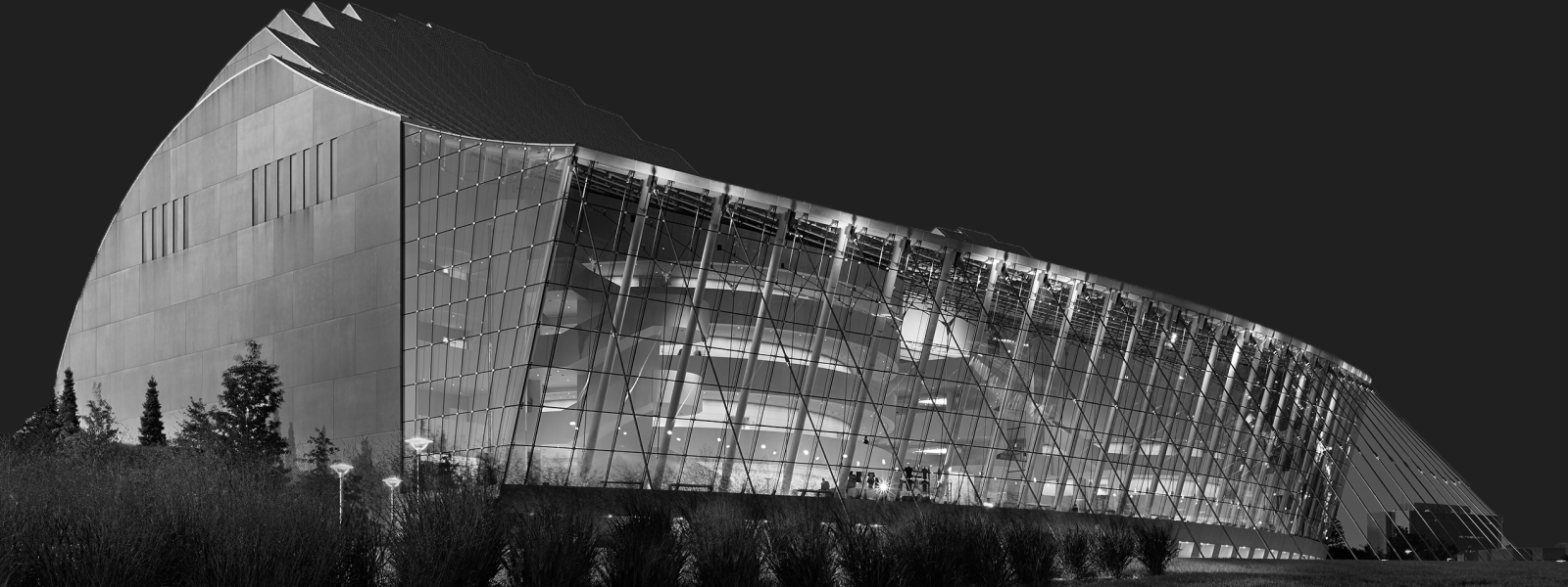 A modern building with a unique geometric design, featuring large glass panels and surrounded by vegetation, illuminated at night.