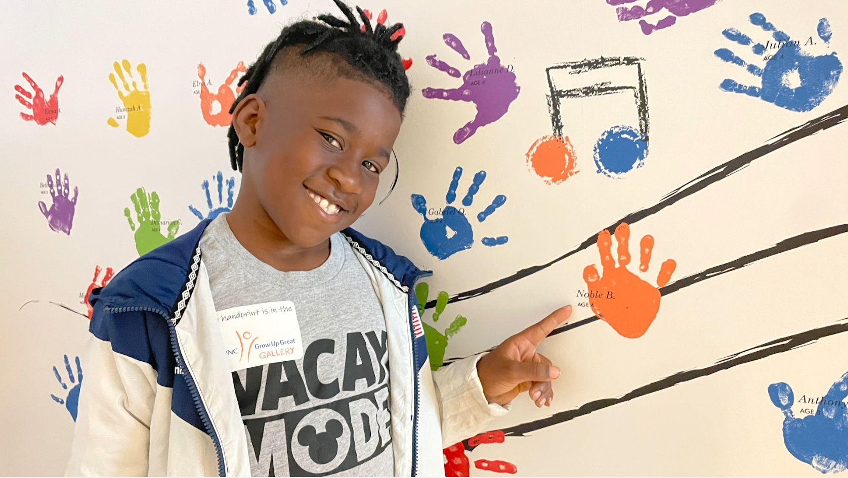 A smiling child points to their orange handprint on a wall covered with colorful handprints and musical note graphics in the PNC Grow Up Great Gallery. The child is wearing a "Vacay Mode" shirt and a jacket.