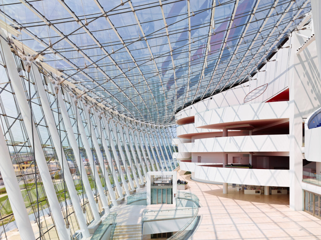 A modern architectural interior with a large, curved glass ceiling, white columns, and multiple levels of open balconies and walkways. The design allows ample natural light into the space.