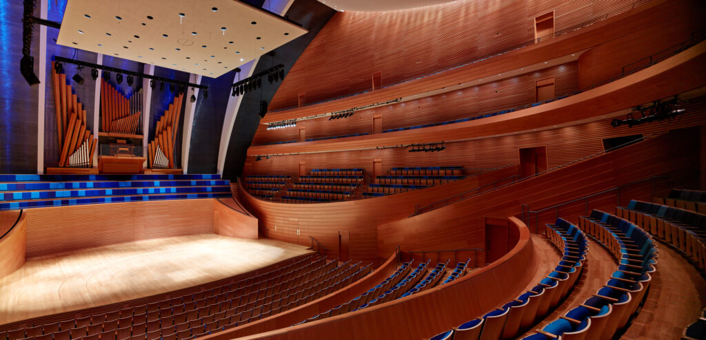 A modern concert hall with wooden interiors, multiple seating levels, and an organ in the background. The stage is brightly lit, and the seating is arranged in curved rows.