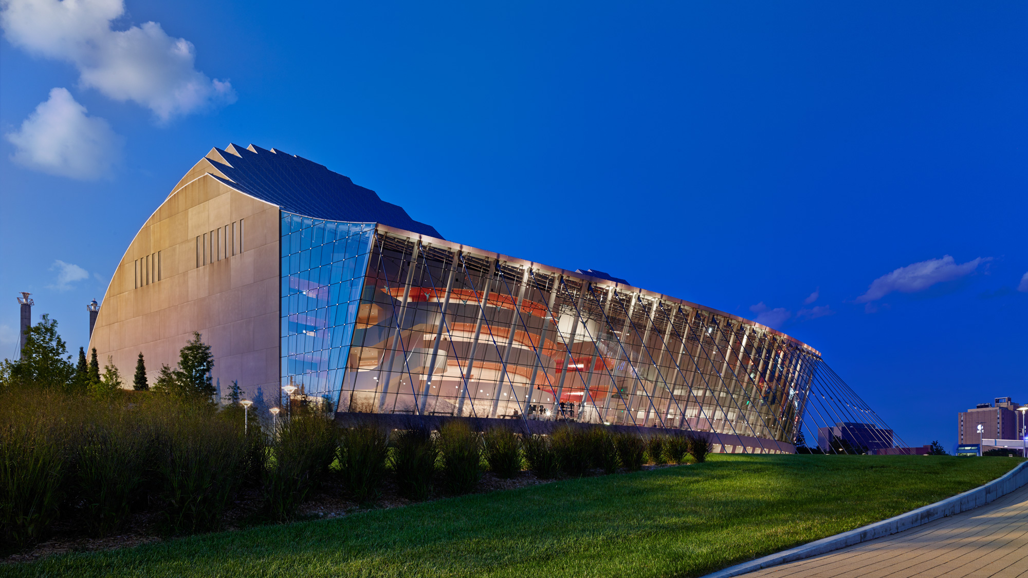 A modern glass-walled building with a curved roof is illuminated at dusk. This stunning example of architecture features large, prominent metal beams and is surrounded by grass, all set against a backdrop of clear blue sky.