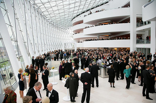 A large crowd of people in formal attire gathers in a modern, open space with glass walls and multiple levels at the Kansas City Performing Arts Center, engaging in conversation and mingling around a central bar area.