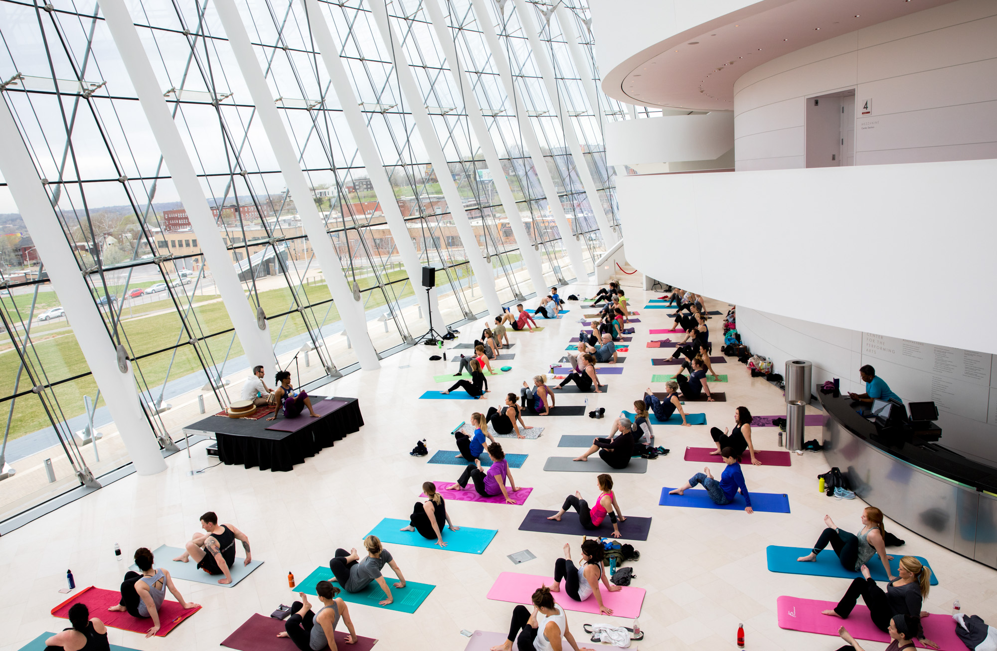 A large group of people participate in a yoga class, practicing various poses on colorful mats in a spacious, modern room with floor-to-ceiling windows and white walls at the Kauffman Center.