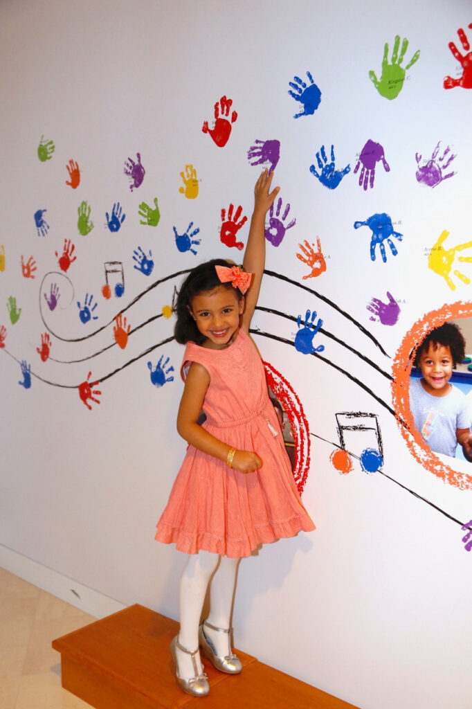 A young girl in a pink dress smiles and points to a purple handprint on a wall decorated with colorful handprints and musical notes at the PNC Grow Up Great Gallery. She stands on a wooden platform.
