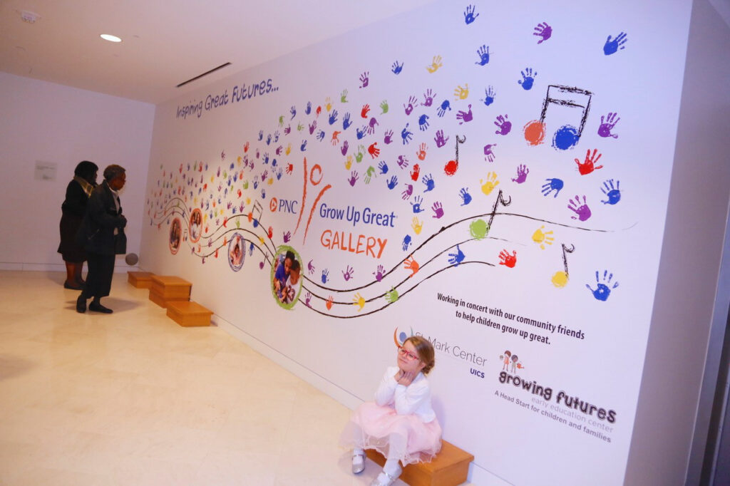 A child sits on a step in front of a colorful mural with handprints and musical notes at the PNC Grow Up Great Gallery. Two adults view the mural. The text includes "Helping Create Futures" and "Growing futures.