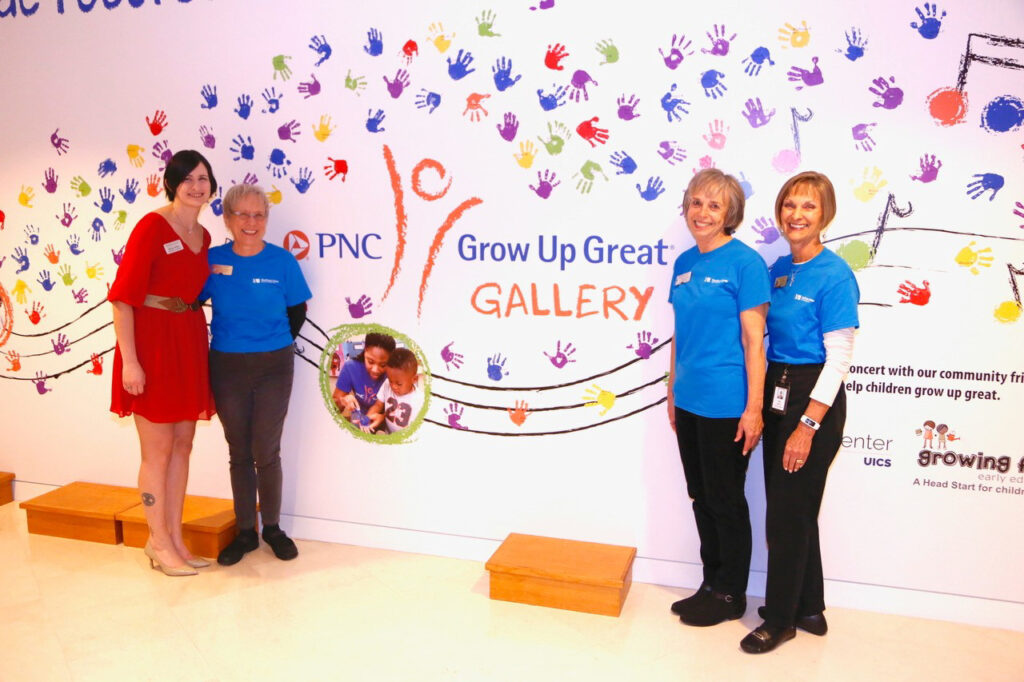 Four women stand in front of a colorful wall with handprints and the "Grow Up Great Gallery" logo. Three are in blue shirts and one in red. The wall includes various sponsor logos.